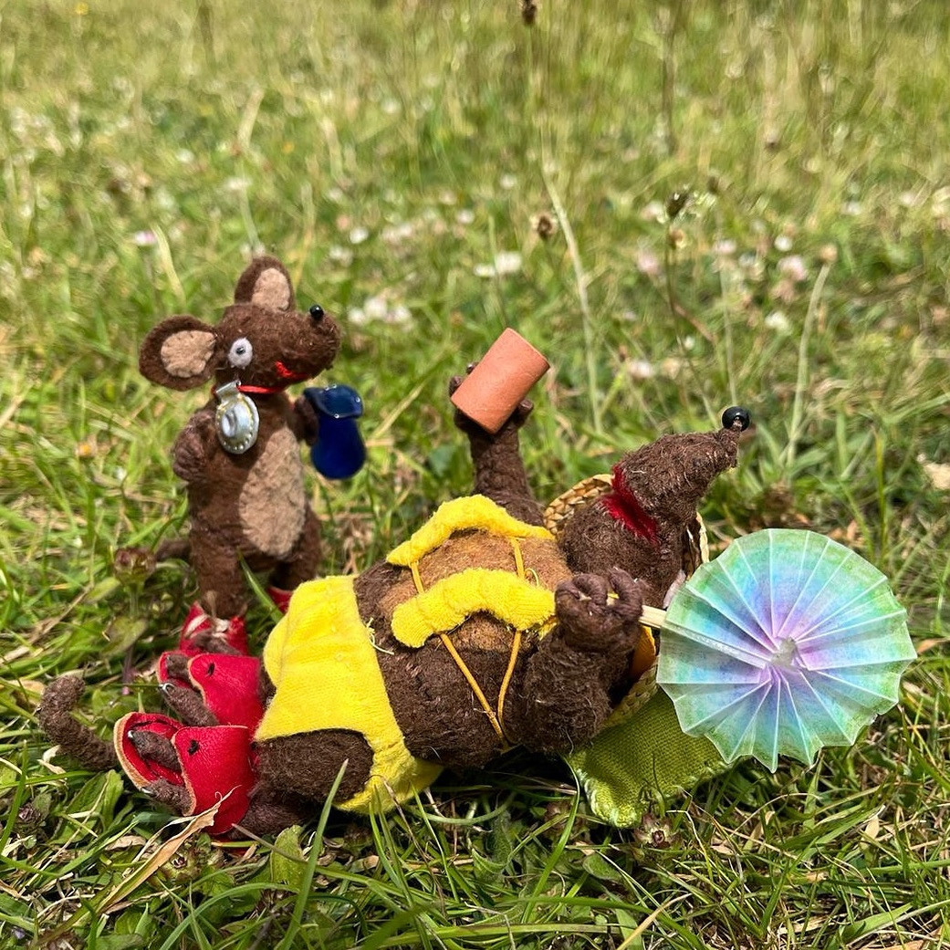 Photo of Minima and Silvius, the Latin mice, in a sunny garden. Minima is wearing a dekakini - a mouse-style bikini with ten cups instead of two, and waving a circular fan. Silvius is kindly bringing a jug to refill her drink.